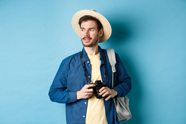 胡子戴着夏帽的帅哥 拿着相机往旁边看 旅游者在度假时拍照 背着蓝色背景的背包站着朋友男休闲