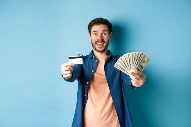 发型开朗的白种人手里拿着美元和塑料信用卡 站在蓝色的背景上 对着镜头开心地笑着年轻自信情感