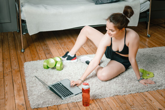 客厅特写镜头中 一位身穿运动服的年轻女子躺在家里的地板上 通过笔记本电脑（一个社交网络）观看电影和学习放松健康运动