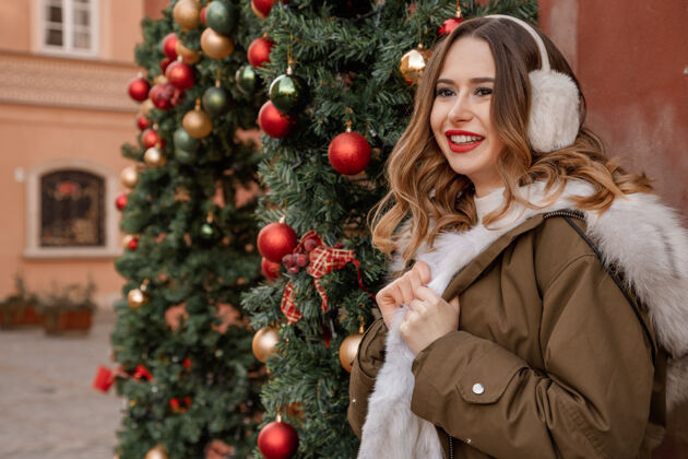 女士穿着针织毛衣和蓬松耳机的女士户外写真年轻漂亮快乐的微笑女孩穿着皮草外套在装饰圣诞树附近女性时尚生活方式