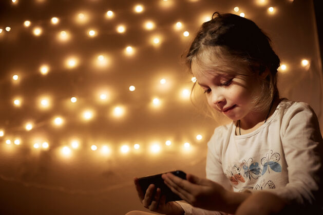 学习小女孩在房间里玩智能手机游戏 一个女孩的画像在一个黑暗的房间里 戴着花环 手里拿着电话无线技术水平家庭内部