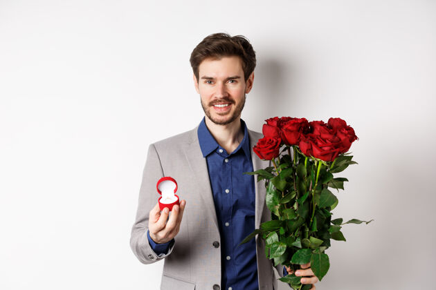 套装微笑的男朋友求婚 手持订婚戒指和一束红玫瑰 浪漫的情人节约会 白色背景玫瑰男人戒指