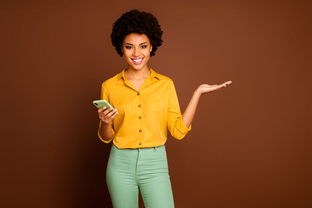 举行积极开朗的非裔美国女孩肖像使用智能手机牵手呈现在线广告促销社交媒体销售穿黄绿色套装隔离棕色皮肤多国混合