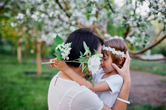童年可爱的女儿和妈妈拥抱在花开的春天花园里快乐的女人和孩子 穿着白色的衣服在户外 春天的季节来了母亲节假期概念女儿花束拥抱