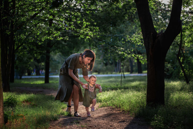 童年女婴迈出了第一步 一位年轻的母亲牵着一个孩子的手 在绿树和树林中帮助她沿着公园的小路行走草.亲本小心女孩支持生活方式