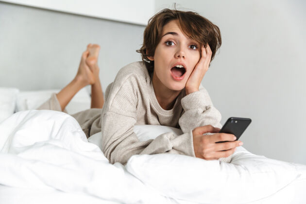 舒适微笑的年轻女孩 早上在床上放松 用手机智能小玩意连接