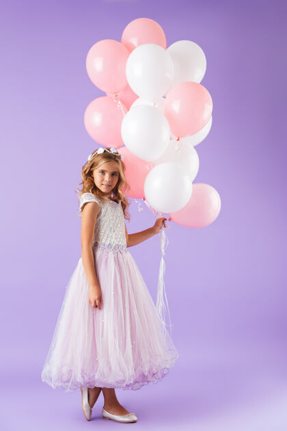 微笑一个穿着公主裙的漂亮小女孩孤零零地站在紫罗兰色的墙上 手里拿着一堆气球快乐女儿吸引力