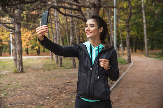 运动微笑的健身女士戴着耳机听音乐 站在公园里自拍户外户外运动员
