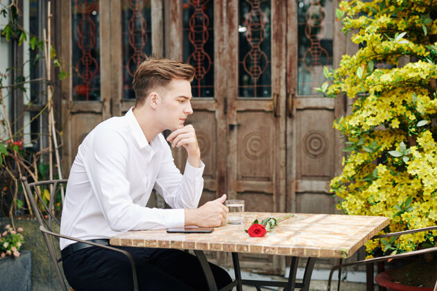 坐一个忧郁的年轻帅哥坐在户外咖啡馆的桌子旁 看着红玫瑰送给他的女朋友男人男性美食物