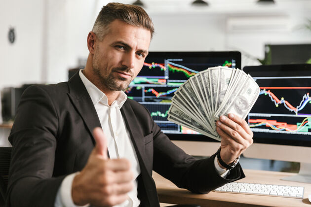 白种人图为30多岁的成功商人穿着西装拿着钱扇在办公室工作 电脑上有图形和图表金钱导师现金