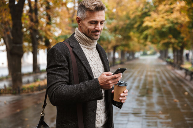 休闲图为30多岁的时尚男子汉穿着暖和的衣服在户外穿过秋天的公园 并使用手机空旷夹克时尚