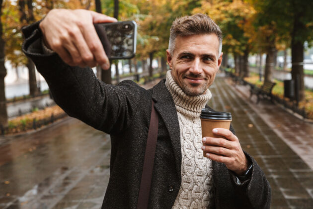 胡须30多岁的成熟男人穿着暖和的衣服在户外穿过秋园 用手机自拍的照片成人肌肉白种人