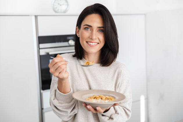 吃一个30多岁的黑发女人站在家里的现代厨房里吃玉米片当早餐成熟燕麦片白种人