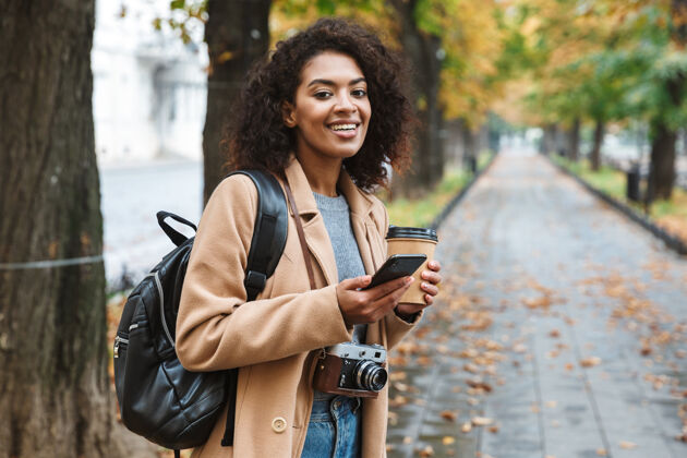 电话快乐的非洲年轻女子穿着外套 拿着外卖咖啡杯走在户外年轻手机青少年