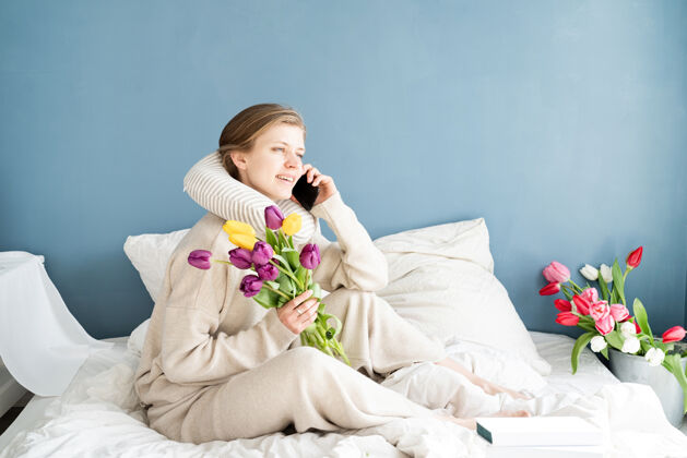 笔记本电脑快乐的女人穿着睡衣坐在床上讲电话 蓝色的墙壁背景交谈笑电话