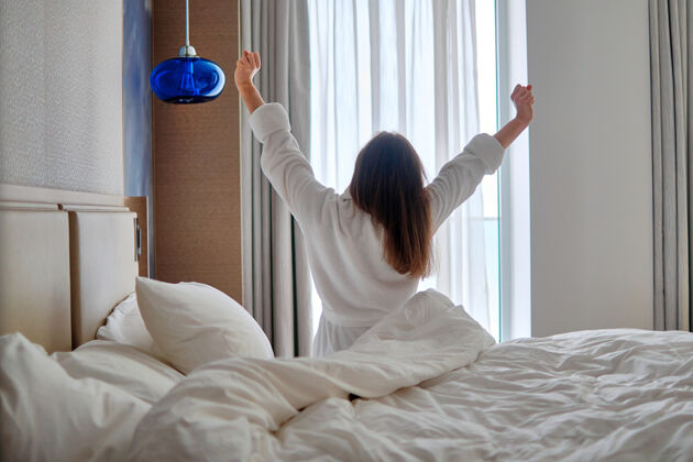 早晨后视图穿着浴衣的懒女人在酒店房间舒适的卧室里放松时 清晨从良好的睡眠中醒来懒散唤醒回来