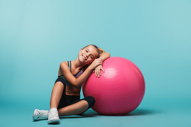 童年快乐的小女孩穿着运动服 靠在一个健身球上 隔着蓝色的墙幼儿园健身球孩子