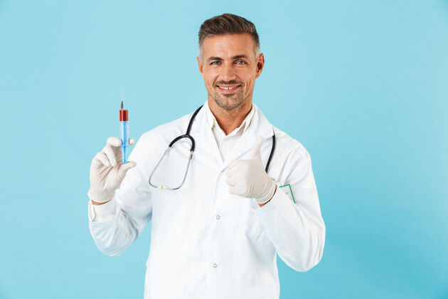 科学自信的男医生穿着制服 孤立地站在蓝色的墙上 展示着一个装有红色液体的注射器医学艾滋病白种人