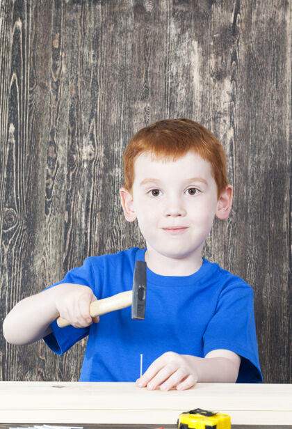 外观红发男孩的欧洲外观在建造一个木结构 在他手里拿着锤子和钉子工作工具修理
