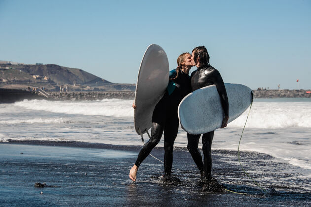 生活快乐的一对穿着黑色潜水服的冲浪者手持白板 在水里亲吻天空和海洋西班牙女孩快乐