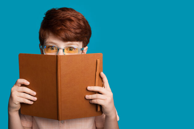 人戴着眼镜的姜男孩躲在一本书后面 看着一个蓝色工作室墙上的摄像头 里面有空想法成功学生