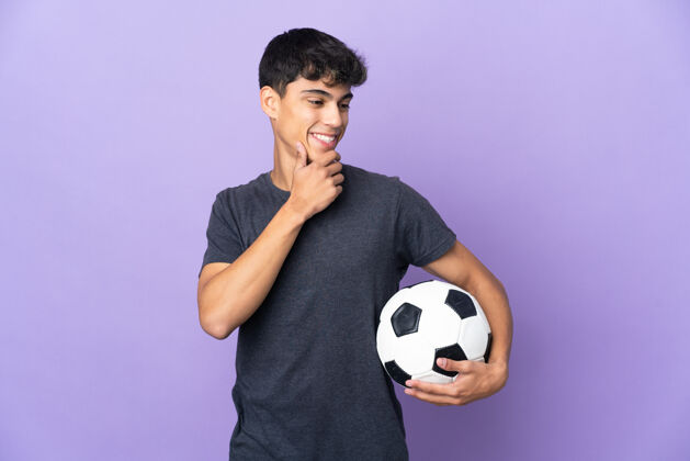 球一个年轻的足球运动员在孤立的紫色背景下看着一边微笑着年轻球员微笑