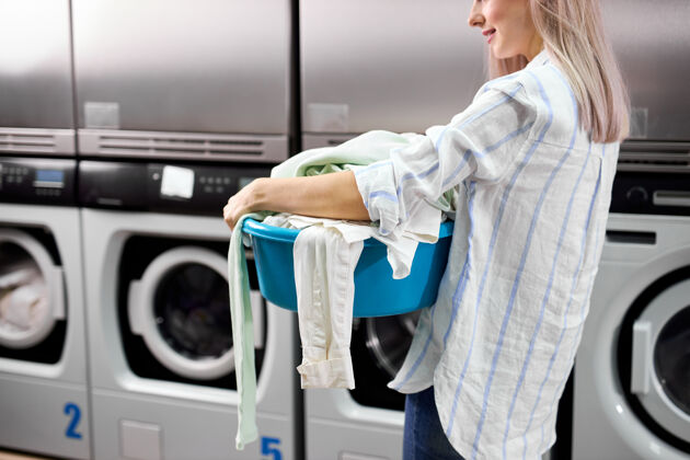 清洁带烘干机的自助洗衣房里 一个衣衫褴褛的女人独自站着 拿着脏衣服机器.内螺纹在休闲装的立场举行盆地与衣服焦点在女人身上人电洗