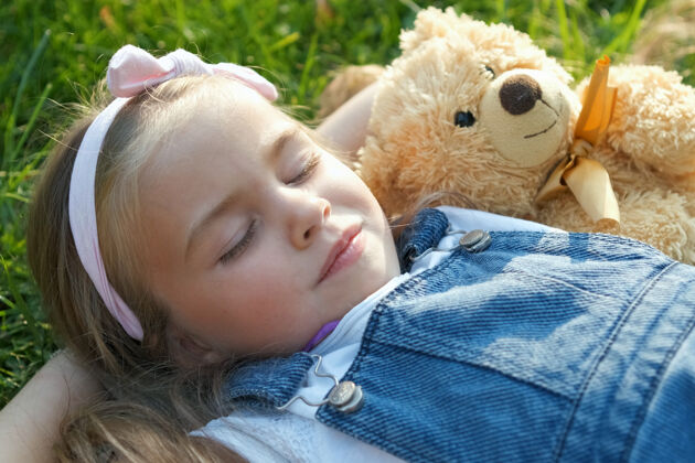 户外美丽的小女孩闭着眼睛躺在毛毯上她的泰迪熊玩具在绿草在夏天午睡熊睡眠下来
