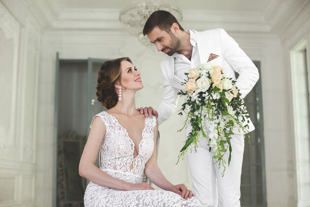 服装别致的婚礼新人新郎和新娘在一个白色的姿势工作室快乐夫妻笑怀特连衣裙白色套装 青春 婚礼.吊灯沙发门门拍摄乐趣