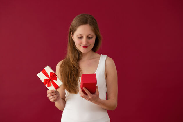 抱着在红色背景上看着礼物的金发女孩快乐微笑盒子