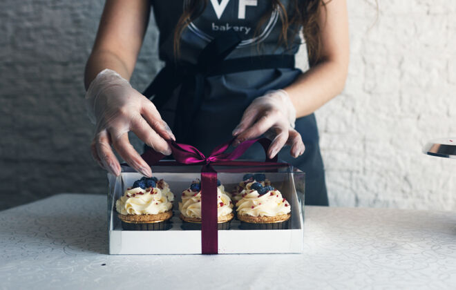 自然一个穿着灰色围裙的女孩在一个礼品盒里包上奶油纸杯蛋糕 把订单送到餐厅客户.烘焙在家里商店纸杯蛋糕厨房