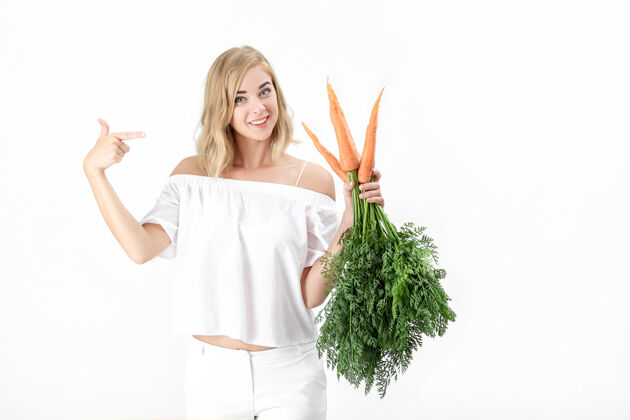 吃一位穿着白色上衣的金发美女在一张白纸上展示了一个绿叶的新鲜胡萝卜背景.健康还有节食女性美丽自然