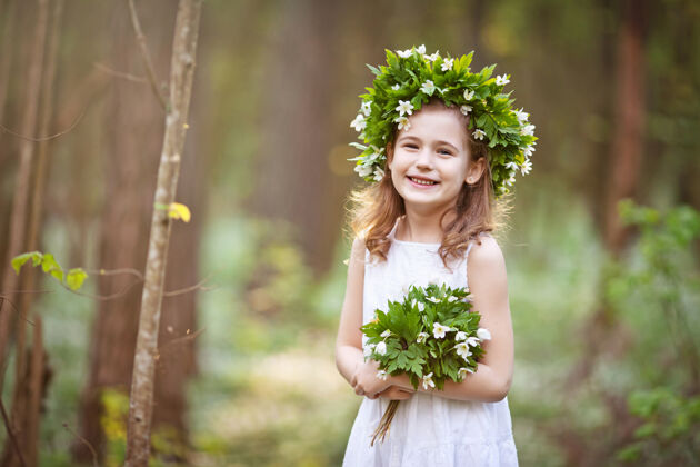小穿着白色连衣裙的美丽小女孩在春天的树林里散步一幅美丽小女孩的肖像 头上戴着春天的花环阳光活动森林