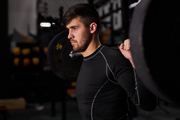 个人运动健将杠铃年轻高加索肌肉发达的男性健美运动员在黑暗的健身房做举重锻炼 使用运动设备 男人站在一边看 一边看年轻动作独自一人