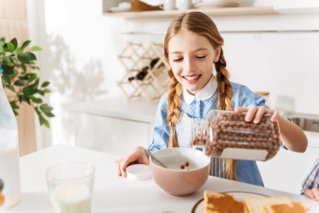 烹饪美味可口甜蜜的愉快的聪明漂亮的孩子在享受阳光明媚的早晨 站在厨房的桌子旁 往碗里装满巧克力片食物厨房吃
