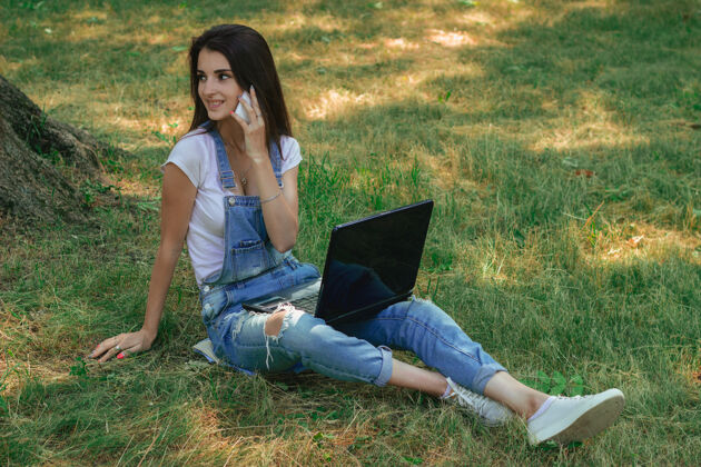 职业一个迷人的年轻女子坐在草地上拿着笔记本电脑和电话服务笔记本电脑活动