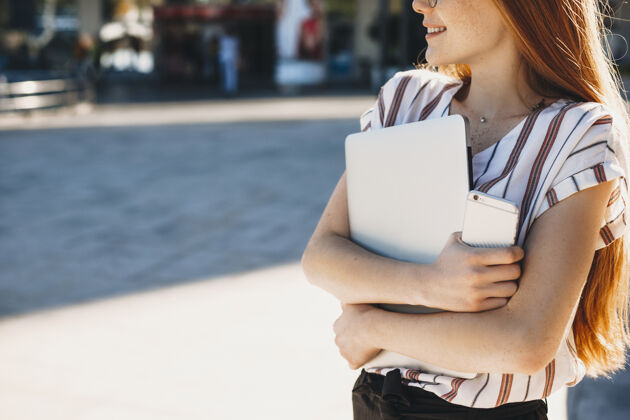 红发特写镜头是一个年轻的女性手拿着笔记本电脑和智能手机在外面对着一栋楼摩天大楼学生街道