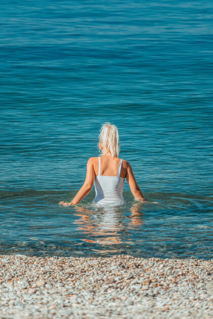 成人后视图：苗条的年轻棕褐色女士穿着白色泳衣 准备游泳 一半站在清澈湛蓝的水中漂亮女孩长