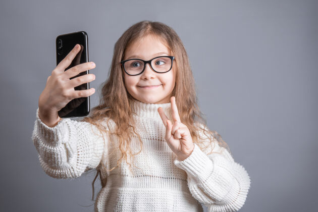 科技一个年轻漂亮的小女孩的肖像 金发飘逸的长发 穿着白色毛衣 手持手机 在视频通讯中自拍可爱自我手机