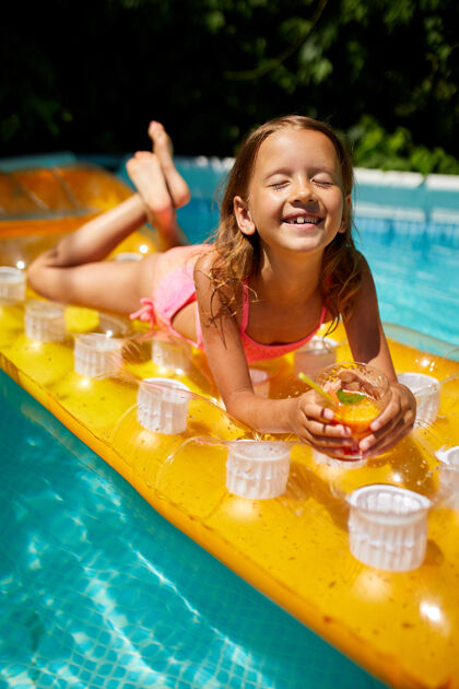果汁小女孩在游泳池里放松 享受日光浴 在家庭度假时在水里的黄色充气床垫上喝果汁度假欢呼夏天