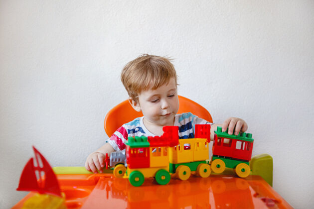 活动一个金发小男孩坐在家里一张橙色的儿童桌旁 玩着五颜六色的塑料玩具套件高高质量的照片积木房子金发