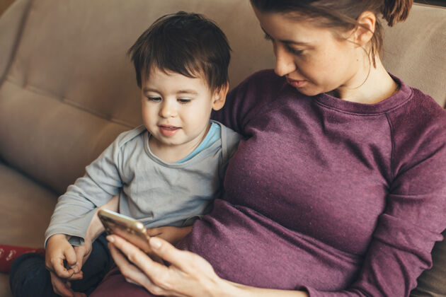 父母怀孕的白人妇女和她的小儿子坐在沙发上看电话等待儿子应用程序