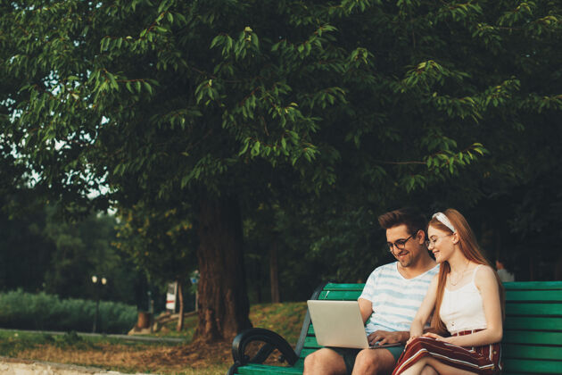 男朋友可爱的年轻夫妇坐在公园的长椅上看着笔记本电脑对着树笑朋友社交肖像