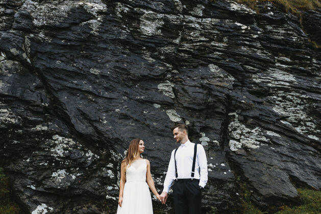 妻子可爱的新郎和新娘手牵着手 面带微笑地看着山上的一块黑色岩石山丘男性浪漫