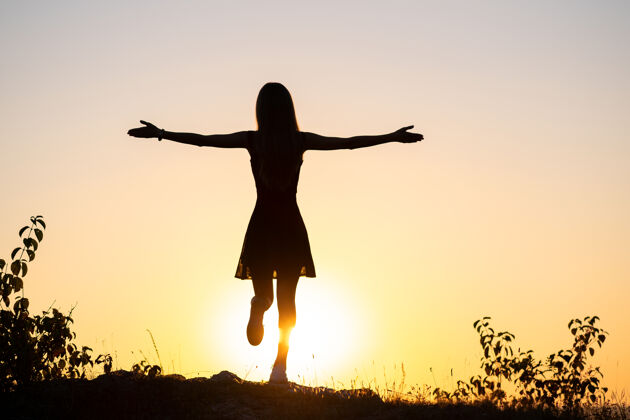 宁静一个年轻女子举起双手站在石头上欣赏夏日户外日落景色的黑色剪影和谐放松能量