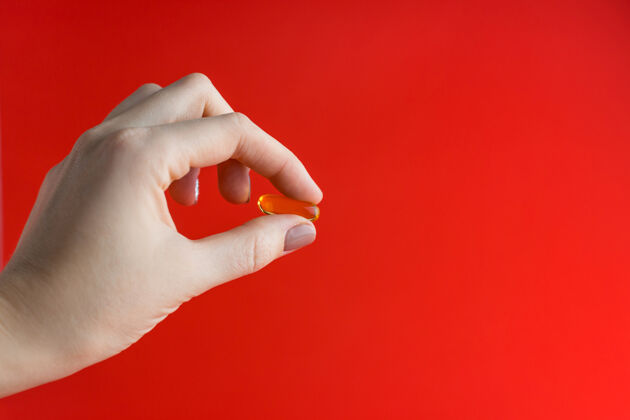 治疗黄色药丸夹在两个手指之间一个女孩的手拿着一个红色的透明药丸背景.金色鱼油胶囊 雌性棕榈中的欧米茄-3鱼油治疗药房黄色