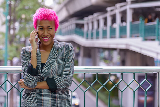 交谈年轻叛逆的亚洲女商人的肖像 粉红色的头发在城市户外的人行天桥上风格发型手机