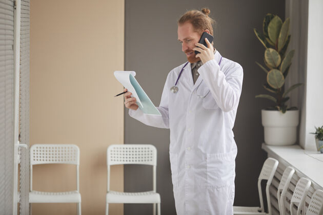 外科医生一位身穿白大褂的男医生站在医院的走廊上 一边看医疗卡 一边用手机咨询自信手持医学职业