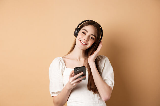 欢呼可爱的微笑女孩戴着无线耳机听音乐 用智能手机 站在米色的椅子上科技促销感觉