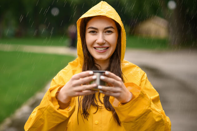 雨穿着雨衣的女人端着一杯热茶 在雨天的夏日公园里 透过窗外的景色一个人女人穿着雨衣走在小路上 雨天在巷子里人季节风暴